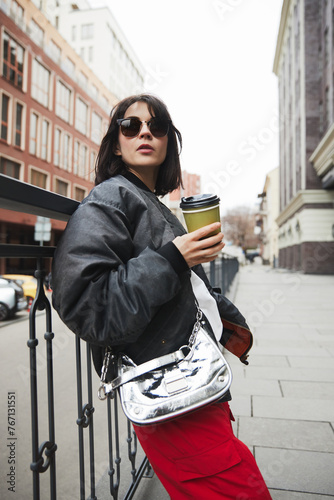 Young woman in stylish gray jacket, metal colored bag and red pants standing with coffee cup, walking around city. Concept of street style fashion, beauty, modern trends