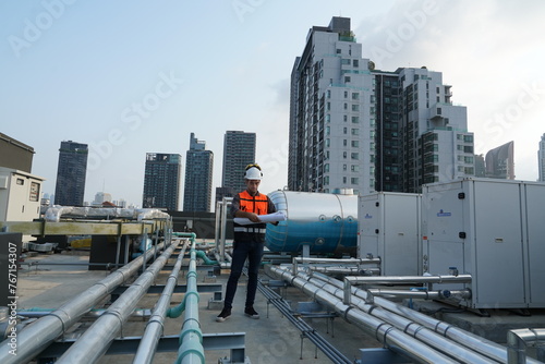 Caucasian male engineer wearing reflective jacket, hard hat holding blueprints inspecting electrical plumbing system on roof top of construction building.