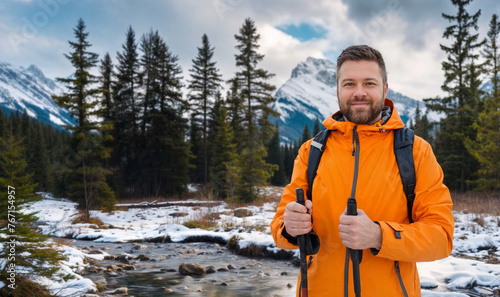 A handsome bearded middle-aged man in an orange traveling jacket holding hiking poles in a Canadian pine forest against the background of a stream and mountains