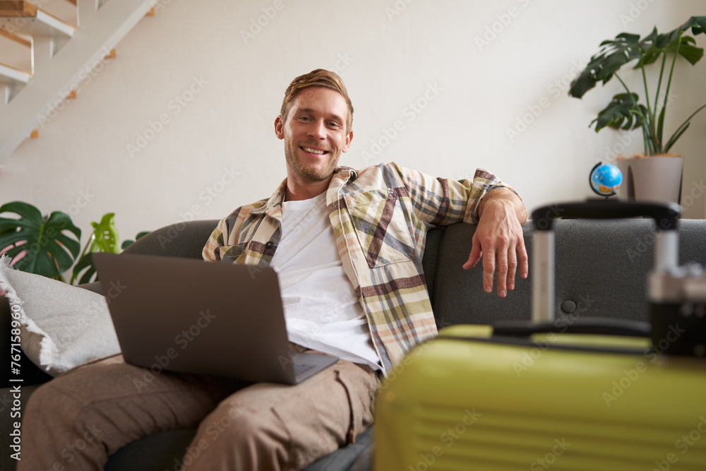Portrait of happy young man, tourist booking his holiday with travel agency, looking at laptop, packed suitcase to go on vacation - obrazy, fototapety, plakaty 