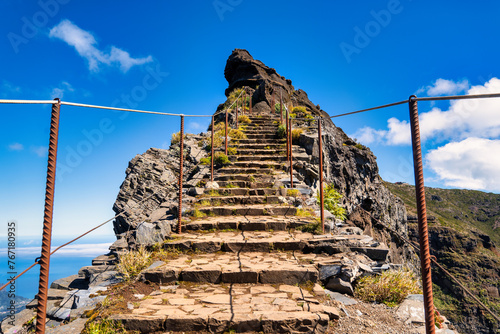 The picture depicts the PR1 Vereda do Areeiro trail in Madeira. A rocky path surrounded by lush greenery leads towards majestic mountain peaks piercing through a sea of clouds under a clear sky.