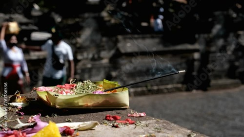 Tripod footage of canang sari with burning incense and Balinese people on the background (out of focus) in Uluwatu temple on sunny day. Bali, Indonesia. photo