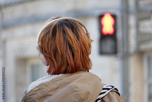 Girl with headphones standing on red traffic light background. Headset, listening to music and city life concept