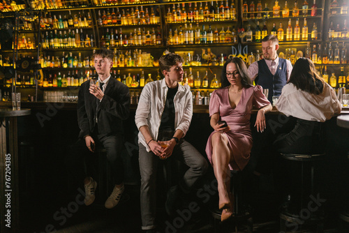 Visitors enjoy cocktails while lady sits lonely nearby in fancy club. Young woman responds to messages on smartphone at night party