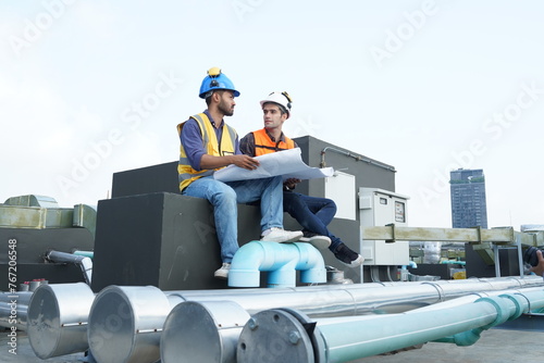Foreman engineer and contractor engineer wearing reflective jacket, engineering helmet, holding blueprints, sit and talk about construction plans on the roof of a building under construction 