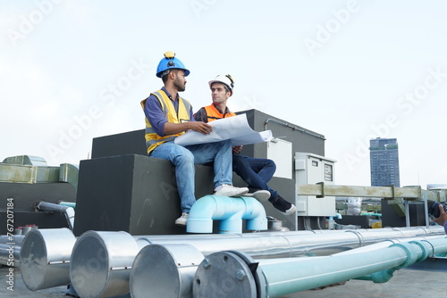 Foreman engineer and contractor worker wearing reflective jacket, helmet, resting from building insurance maintenance inspection on construction building rooftop. 