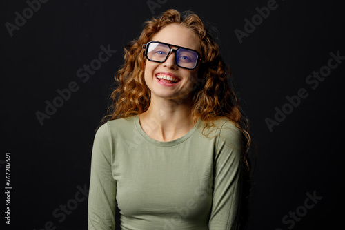 Happy smiling curly young woman wearing glasses looking at camera with joyful smile at black background
