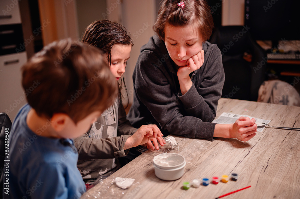 An absorbed group of siblings conduct a science experiment at home under the watchful eye of a young woman, fostering a collaborative and educational family moment