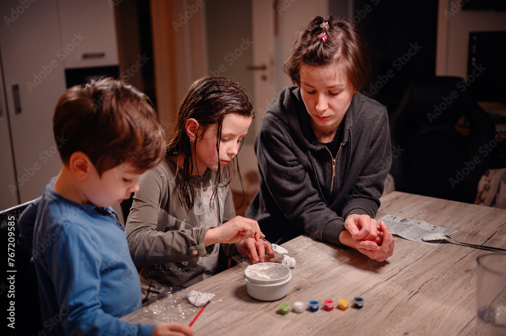 An absorbed group of siblings conduct a science experiment at home under the watchful eye of a young woman, fostering a collaborative and educational family moment
