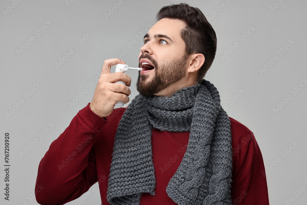 Young man with scarf using throat spray on grey background - obrazy, fototapety, plakaty 