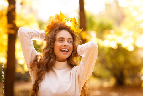 Smiling young woman with the fall crown of yellow leaves on her curly hairstyle head in the autumn park at sunset