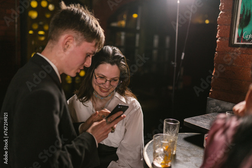 Old friends meet after vacation and discuss different stories. Guy tells girl about abroad trip and shows photos near bar table