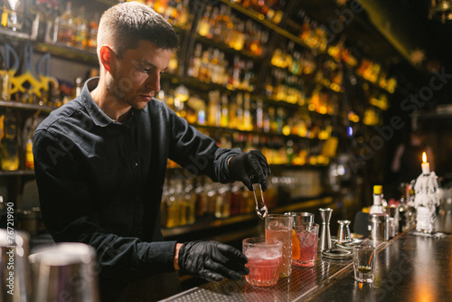 Barman adds ice to pink alcohol cocktail in glass at counter in club. Professional bartender makes delicious beverage in bar