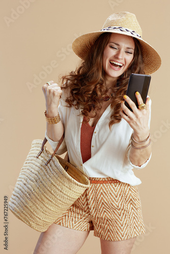 smiling woman using phone with raised arms rejoicing on beige