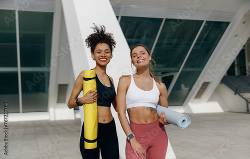 Two smiling female friend standing on morning city background with yoga mats after doing morning gym