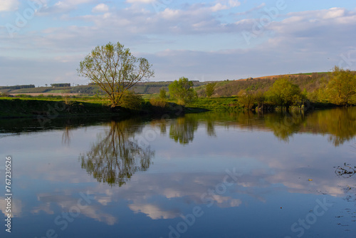 Tranquil landscape at a lake, with the vibrant sky, white clouds and the trees reflected symmetrically in the clean blue water