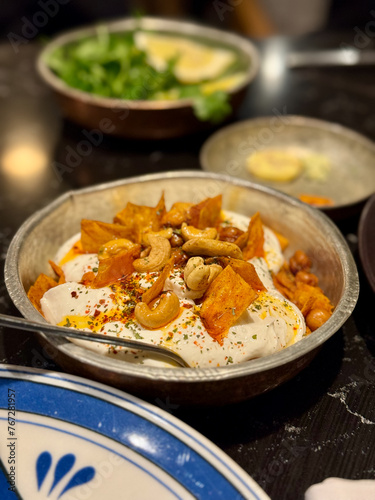 A dish of creamy labneh topped with roasted sweet potatoes, cashews, and seasoning, served in a rustic bowl with a blurred background of greens.