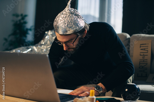 Conspiracy theory. A man in a tinfoil hat looks for signs while watching a video on a laptop.