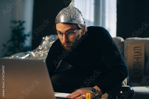 Conspiracy theory. A man in a tinfoil hat looks for signs while watching a video on a laptop.