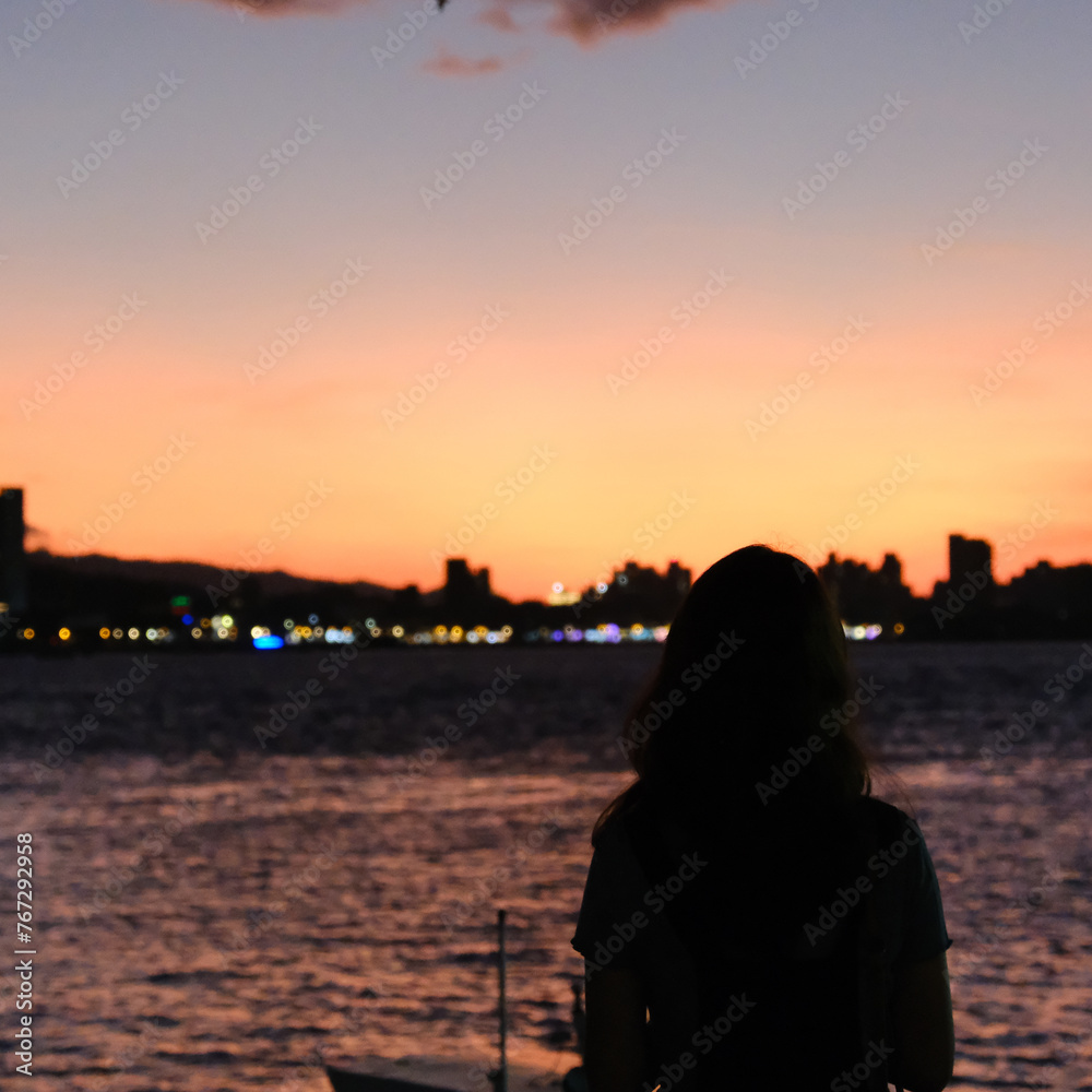 Fototapeta premium Silhouette of a young woman contemplating a vivid sunset over the city skyline at Tamsui Wharf in Taipei, Taiwan.