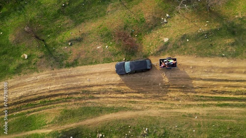 Aerial high angle top down shot of a black car towing a small trailer with two off road cross morotbikes along a windy dirt road. Drone footage of vehicle with a trailer behind going to sports event.
 photo