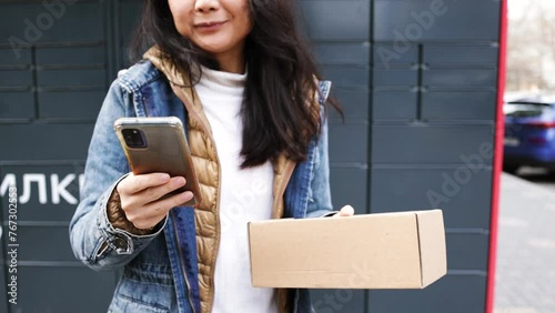 Woman receiving parcel from post terminal machine using smartphone outdoors. Parcel delivery machine. Mail delivery and post service, online shopping, e commerce concept. photo