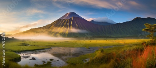 A volcano looms in the background, contrasting with the peaceful river in the foreground. The natural landscape is enhanced by the presence of clouds in the sky and lush grasslands