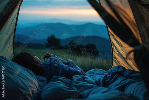  Woman resting in a sleeping bag inside a tent with a mountain view at dusk. Camping relaxation and adventure concept. Design for outdoor gear advertisement, banner, and travel