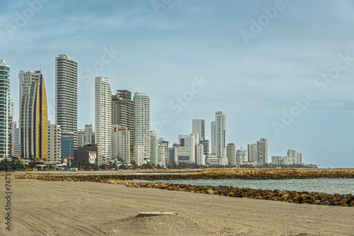 Cartagena, Colombia - July 25, 2023: Entire west coast of Bocagrande peninsula with its tall modern buildings under blue cloudscape. Sandy beach split by rocks from Caribbean sea photo