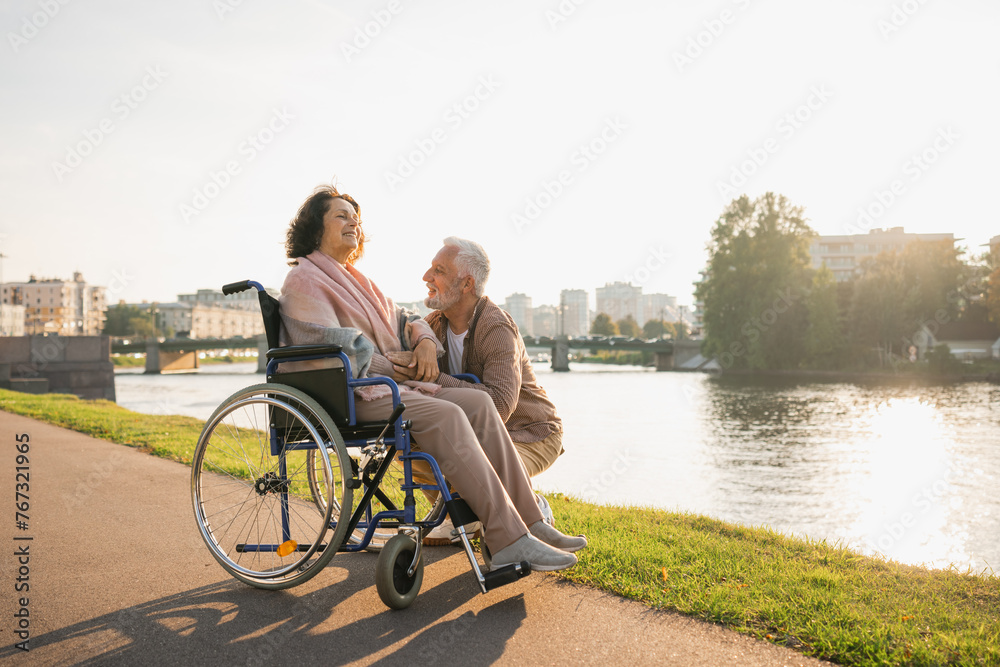 Senior woman in wheelchair walking with caregiver old man on road. Elderly family couple man supporting taking care of paralyzed woman in chair for people with disability outdoor. Rehabilitation Senior woman in wheelchair walking with caregiver old man on road. Elderly family couple man supporting taking care of paralyzed woman in chair for people with disability outdoor. Rehabilitation