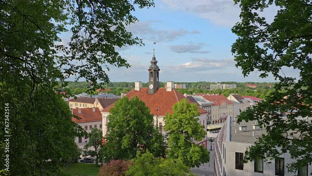 Panoramic view of a classic town hall with a clock tower surrounded by lush trees on a sunny day in Tartu Estonia