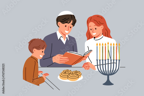Jewish family celebrates day of hanukkah and reads holy book together, standing near table and minors. Jewish mom and dad read encyclopedia to little son, talking about traditions of people of israel