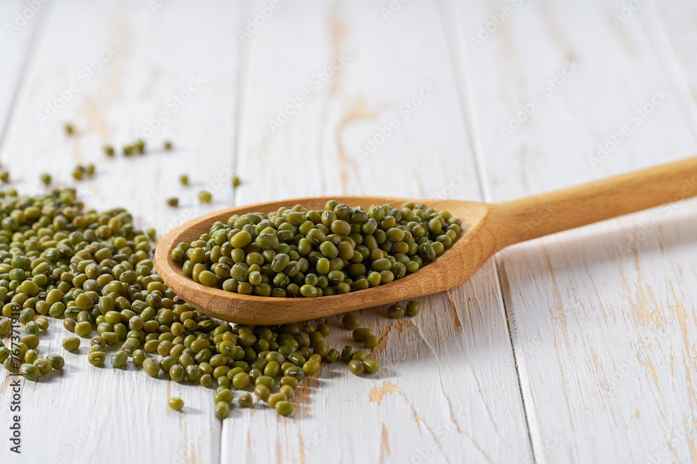 Raw mung beans crumble from in a wooden spoon on a white wooden table. Raw mung beans crumble from in a wooden spoon on a white wooden table.