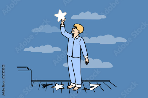 Little boy puts paper star to sky, thinking about endless space and galaxies, dressed in pajamas. Happy child dreams of seeing clean party welcome and wanting to fly into space when grows up