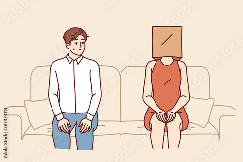 Indecisive man sits near woman with paper bag on head and is afraid of becoming acquainted. Indecisive guy sits on sofa with girlfriend, and is stressed due to timidity interfering with relationships