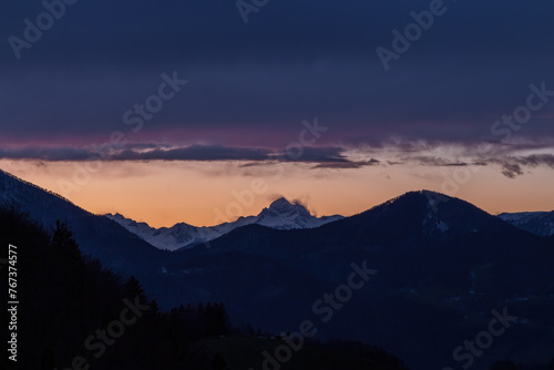 A three peaked mountain in the sunset with cloud cover over them and a small houde in front.