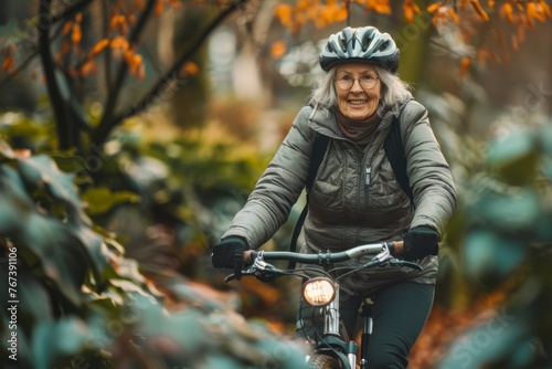 Elderly woman with glasses and helmet cycling through a park with autumn foliage