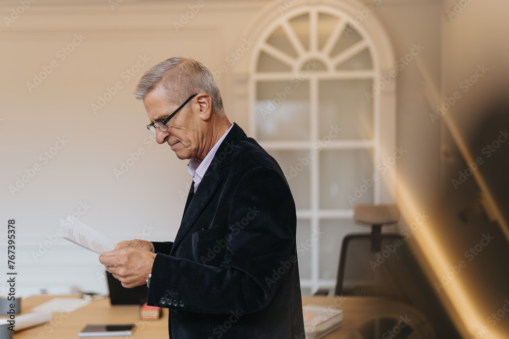 Fototapeta premium Mature businessman reading paperwork, analyzing reports, statistics or teamwork results at modern office setting.