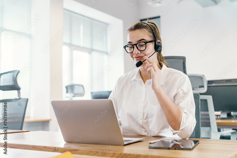 An attentive customer service professional operator is a woman with a headset conducts a call, her gaze fixed on a laptop screen, in a well-lit office. An attentive customer service professional operator is a woman with a headset conducts a call, her gaze fixed on a laptop screen, in a well-lit office.