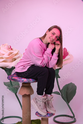 A girl poses with a smile against the background of large artificial white roses. Studio shot with pink lighting