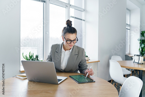 Valokuva A diligent professional woman focuses on her tablet, utilizing a stylus for precision work, with her laptop ready beside her in a minimalist office