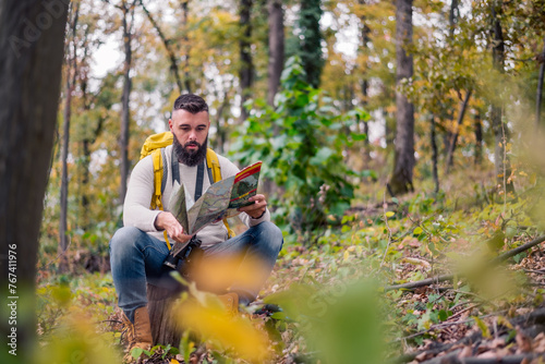 During his woodland hike, a hipster enjoys a rest, sitting on a tree stump while consulting a paper map to plan his route."