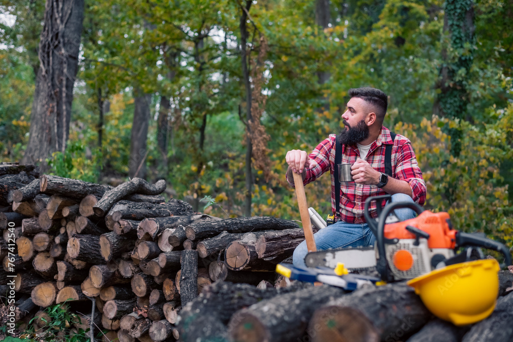 Caucasian man, a lumberjack, takes a moment to rest on a pile of firewood, pouring himself a cup of coffee from a thermos, his tools laid out in front of him. Caucasian man, a lumberjack, takes a moment to rest on a pile of firewood, pouring himself a cup of coffee from a thermos, his tools laid out in front of him.