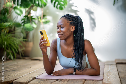 Satisfied relaxed african american woman holding smartphone lying on yoga fitness mat resting after workout. Positive athletic black female looking at screen mobile phone take break in sport training 