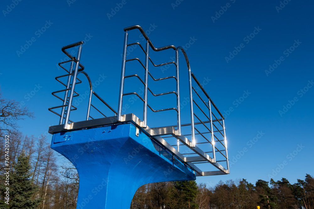 A blue diving board over an empty swimming pool. Stainless steel rails around the platform. The outdoor swimming pool is ready for the seasonal break.