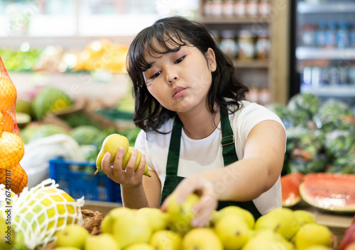 Positive young asian girl working in fruits and vegetables shop  laying out fresh pears on produce display
