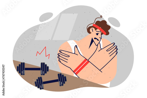 Weak man in gym stands near dumbbells for fitness and holds hands, trying to correct muscle pain