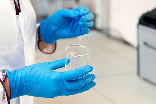 scientist in a white laboratory coat holds in his hands in blue disposable gloves a test tube, glass volumetric measuring cylinder. Working at a desk	