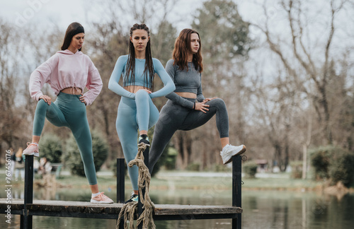 Three fitness enthusiasts take a break for stretching exercises on a jetty over a serene pond, showcasing companionship and an active lifestyle.