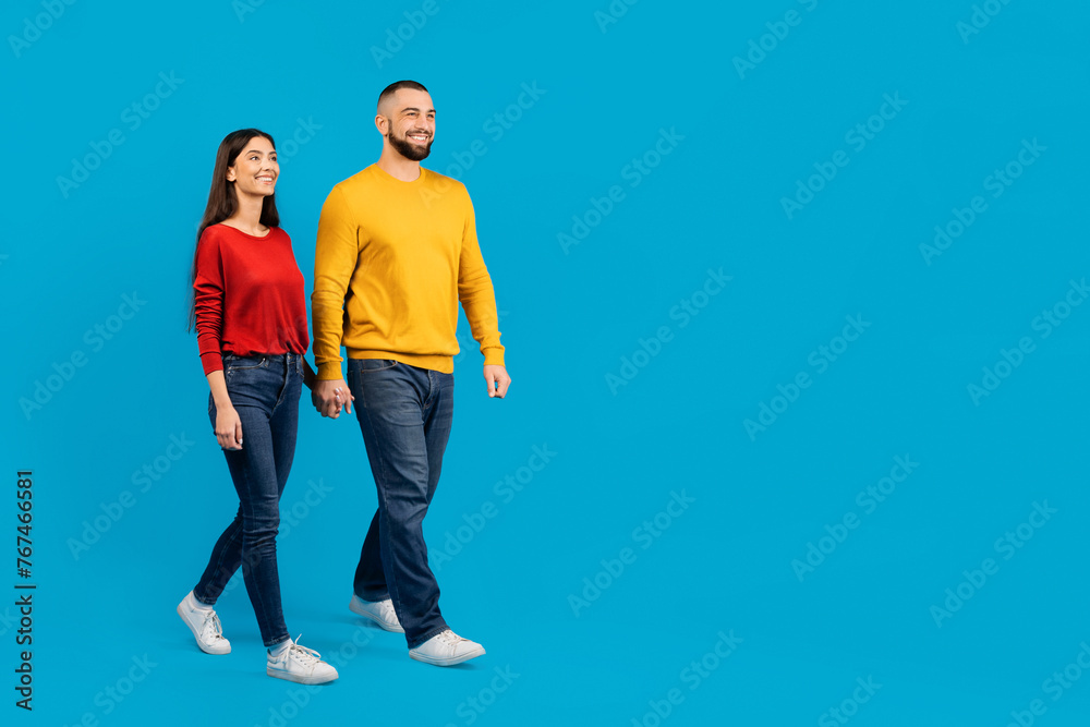 Joyful young couple walking hand-in-hand on blue studio background